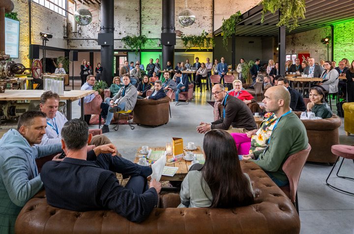 Overzichtsfoto van een informeel werkgeversevent in een industriële, sfeervol ingerichte ruimte, waar groepjes mensen op banken en stoelen bij elkaar zitten, luisterend naar een spreker. Op de achtergrond staan meer deelnemers aan hoge tafels en is er een