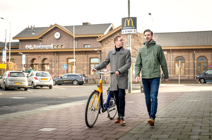 Willie (met fiets) en Xavier wandelen op het stationsplein, op de achtergrond station Roermond